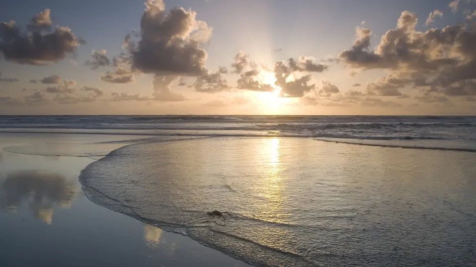Golden sun rays shine through clouds over the ocean at sunset, reflecting on wet sand.