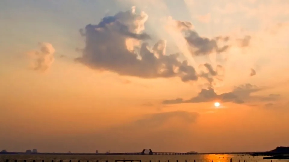 The sun is setting over a body of water, casting a golden glow on the clouds and water.