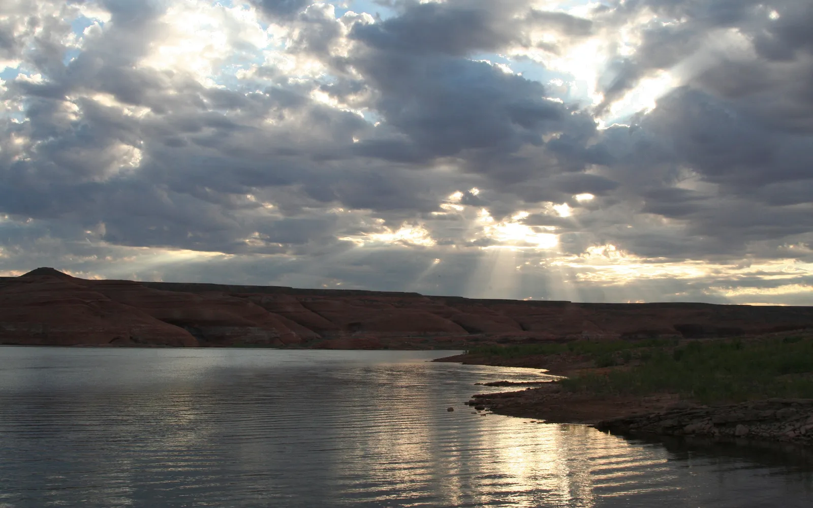 Golden sunbeams break through dramatic clouds over a calm lake with red rock formations in the background.