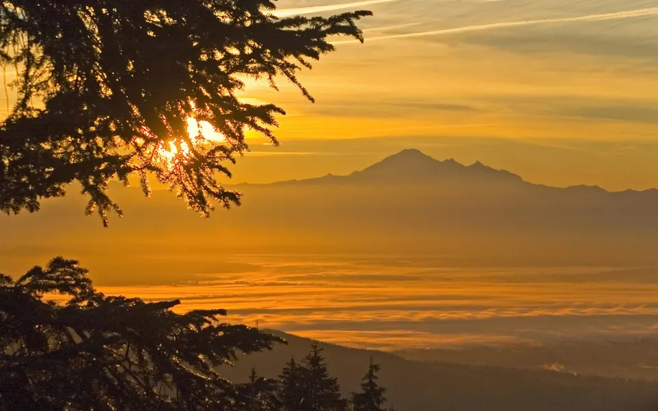 A majestic mountain range is silhouetted against a vibrant orange and yellow sunrise sky, with clouds below.