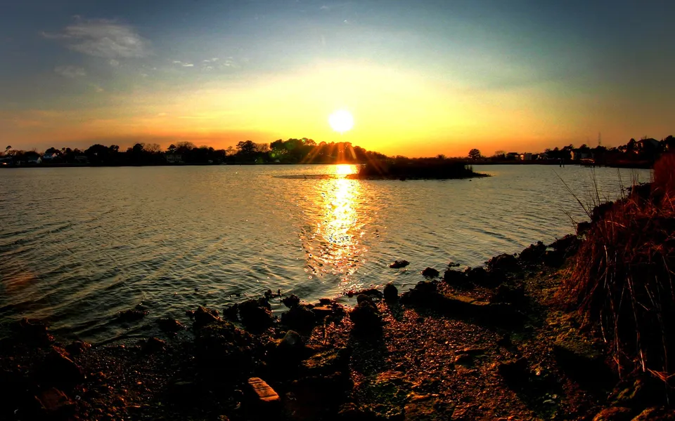 A bright sun sets over a calm body of water, casting a golden reflection on the surface.