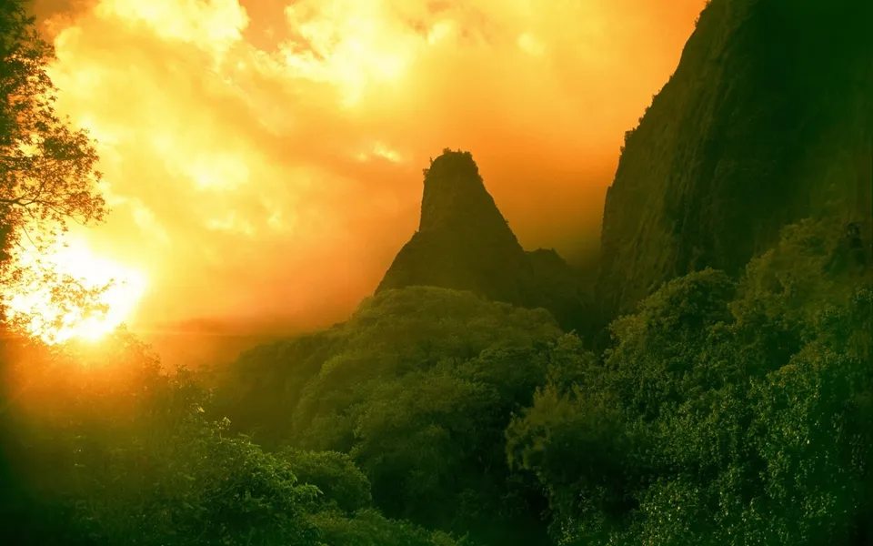 A dramatic sunset casts a golden glow over jagged mountain peaks and a dense green forest.