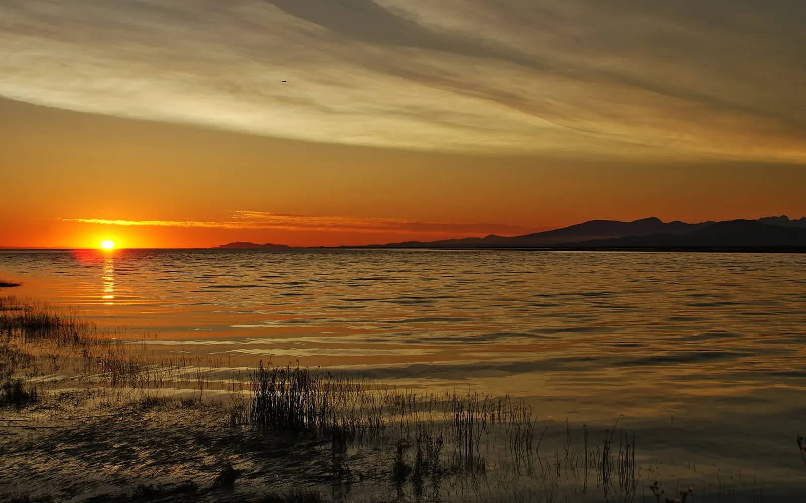 A bright orange sun sets over a calm lake, casting golden light on the water and distant mountains.