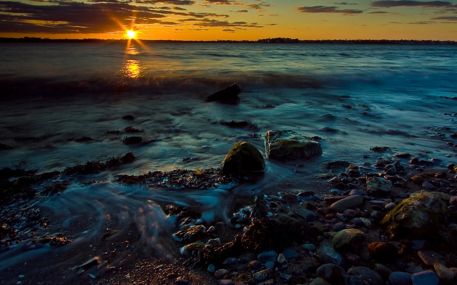 Golden sunset rays reflect on calm water with a rocky shore in the foreground.