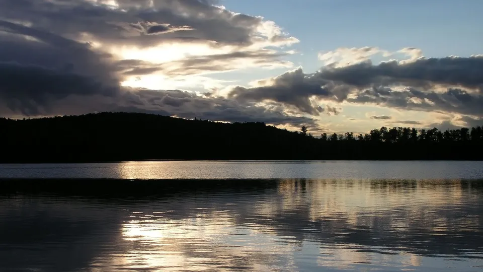 Golden sunset reflection on a calm lake with dark, dramatic clouds and a silhouetted hill.