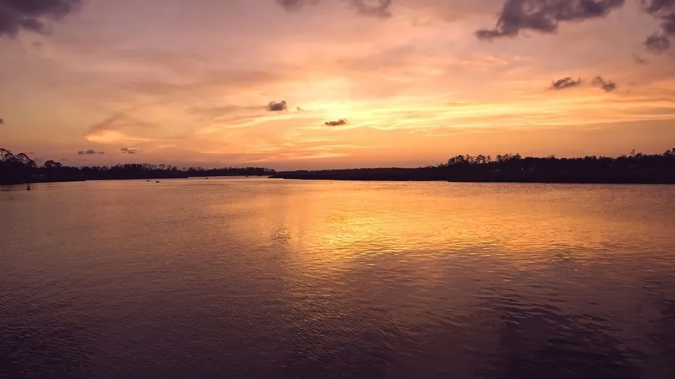 A wide river reflects the warm orange and pink hues of a sunset sky with scattered clouds.