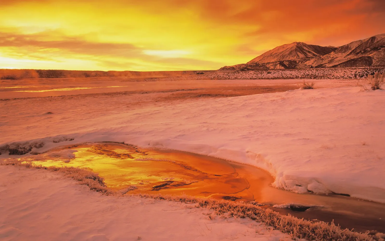 A vibrant sunset casts a golden glow over a snow-covered landscape with mountains in the background and a partially frozen river in the foreground.