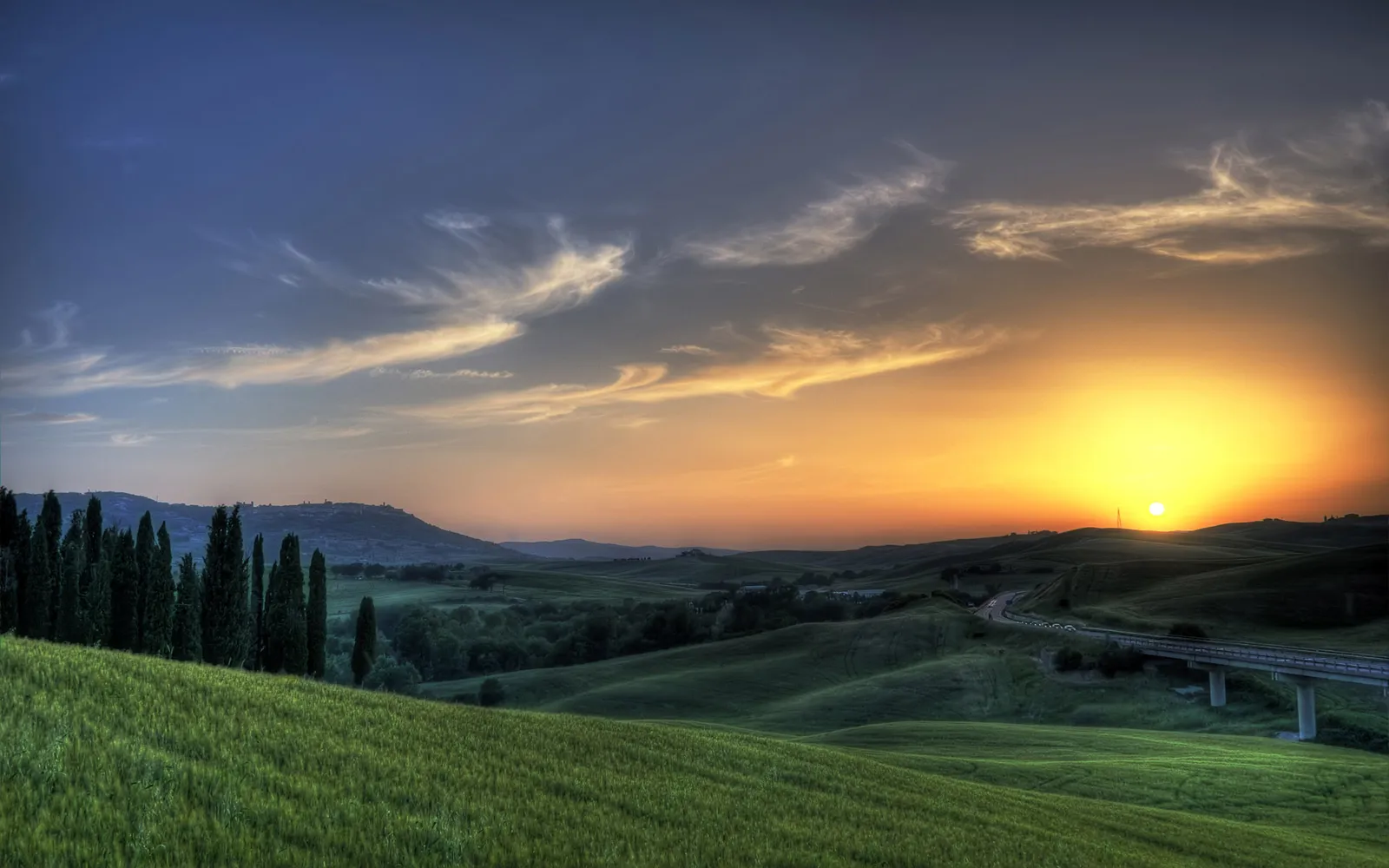 A golden sunset over rolling Tuscan hills, with a line of cypress trees on the left.