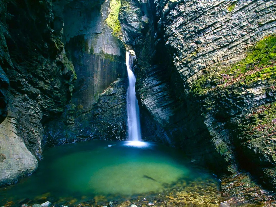 A tall waterfall plunges into a deep green pool surrounded by steep, moss-covered rock walls.