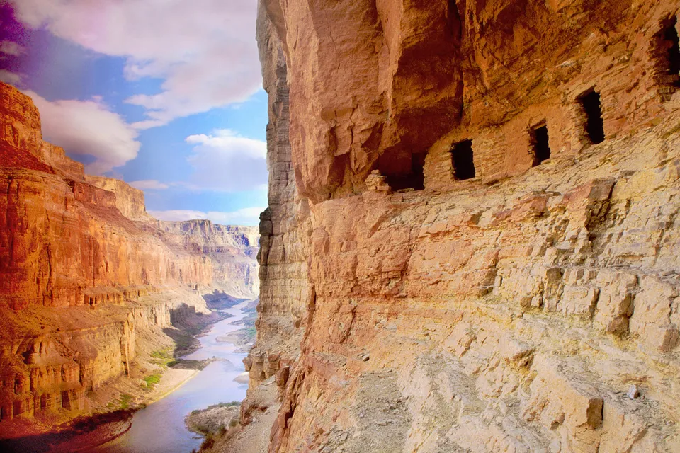 View of ancient stone structures built into the side of a canyon wall, with a river flowing below.