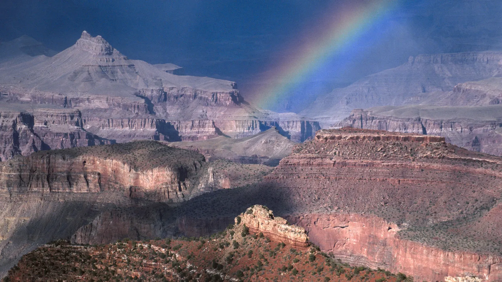 A vibrant rainbow arcs across the vast, layered rock formations of the Grand Canyon under a dark sky.