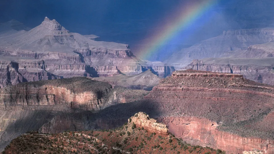 A vibrant rainbow arcs across the vast, layered rock formations of the Grand Canyon under a dark sky.