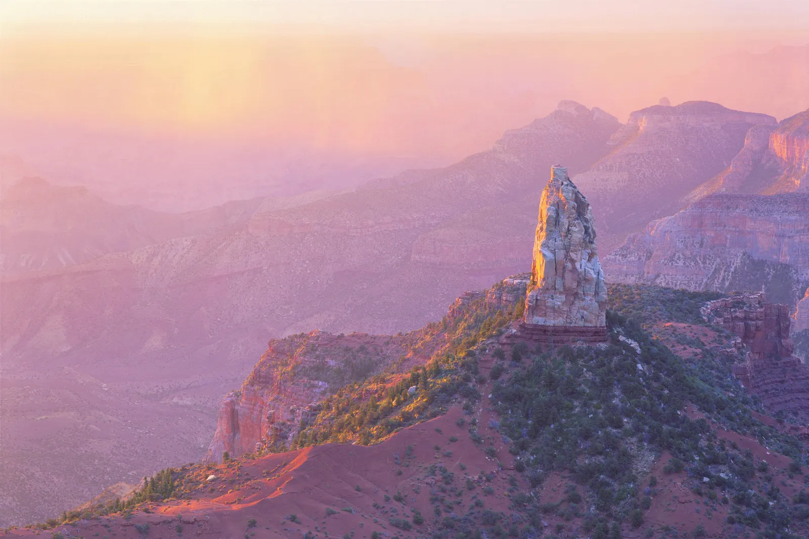 A tall, weathered rock spire stands prominently in the Grand Canyon, bathed in the soft, pink and orange light of sunrise.