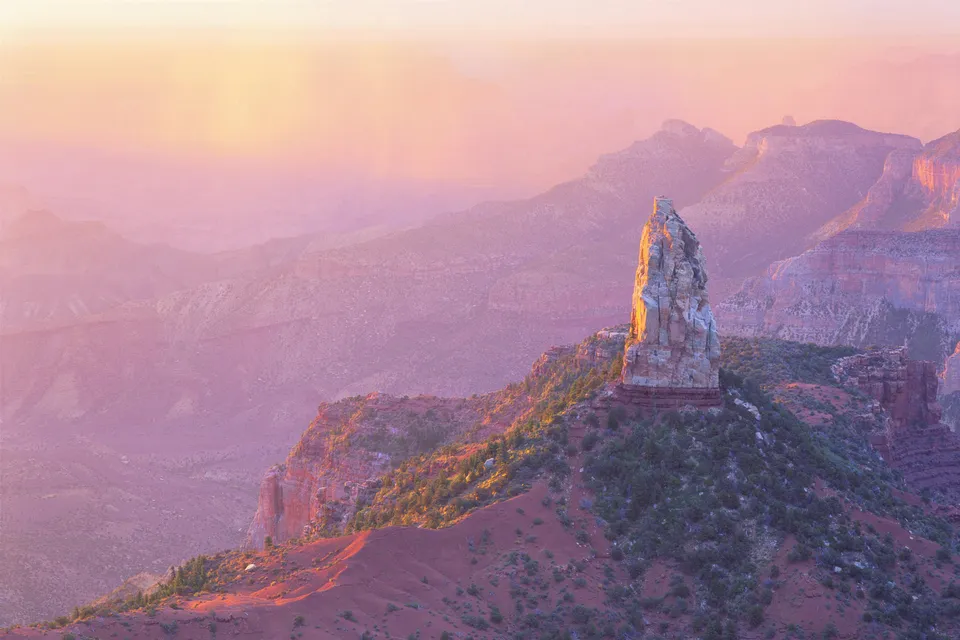 A tall, weathered rock spire stands prominently in the Grand Canyon, bathed in the soft, pink and orange light of sunrise.