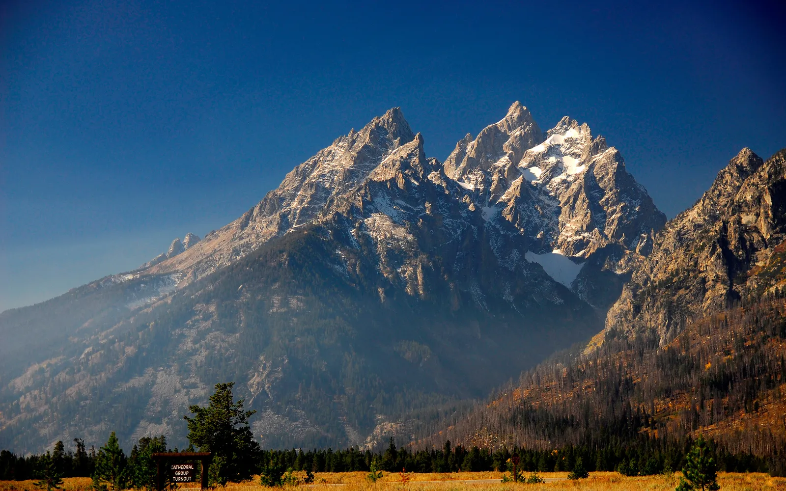 The jagged peaks of the Grand Teton mountain range are illuminated by sunlight against a deep blue sky.