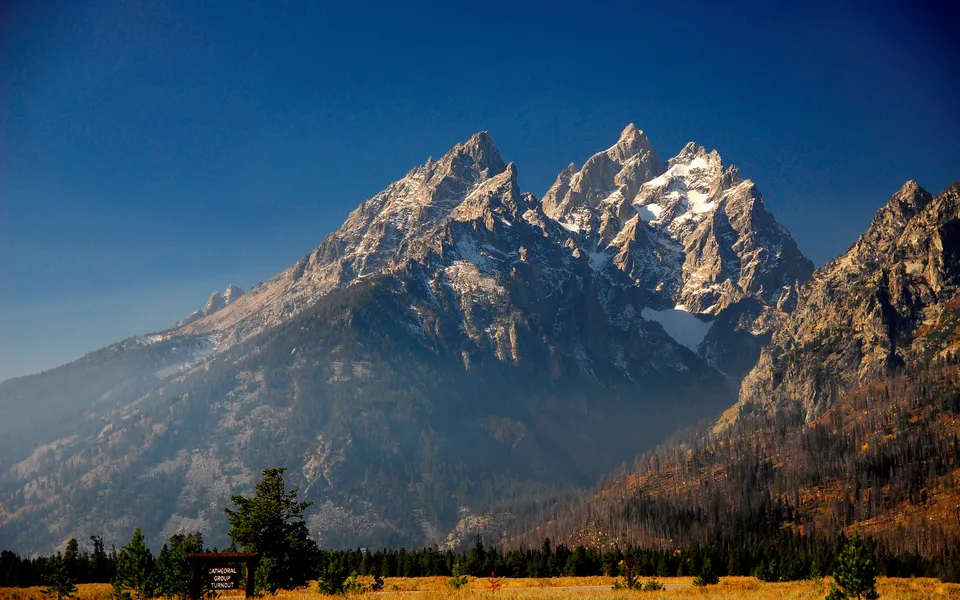The jagged peaks of the Grand Teton mountain range are illuminated by sunlight against a deep blue sky.
