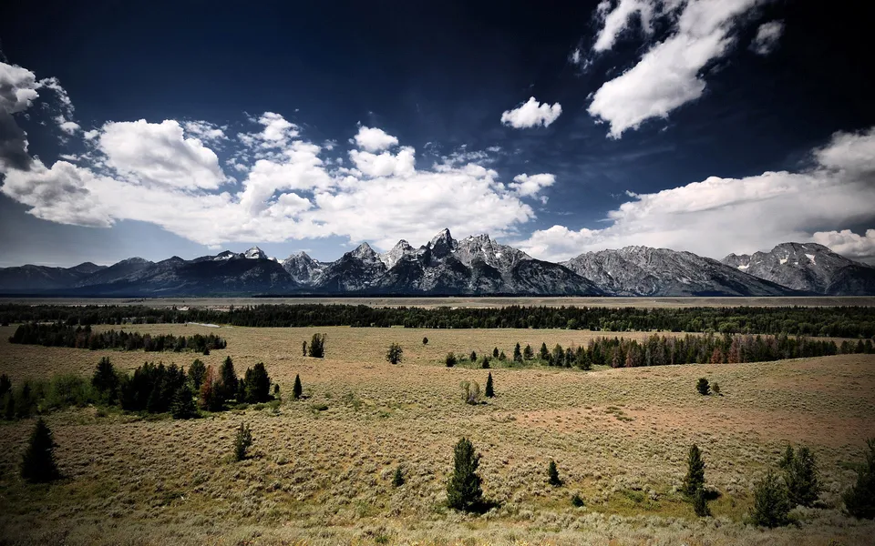 A wide view of the Grand Teton mountain range under a dark blue sky with white clouds.