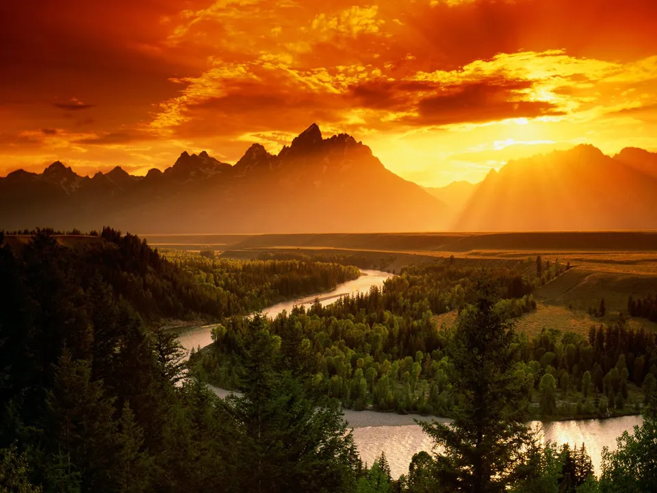A dramatic sunset casts golden light over a winding river and forested valley, with jagged mountains in the background.