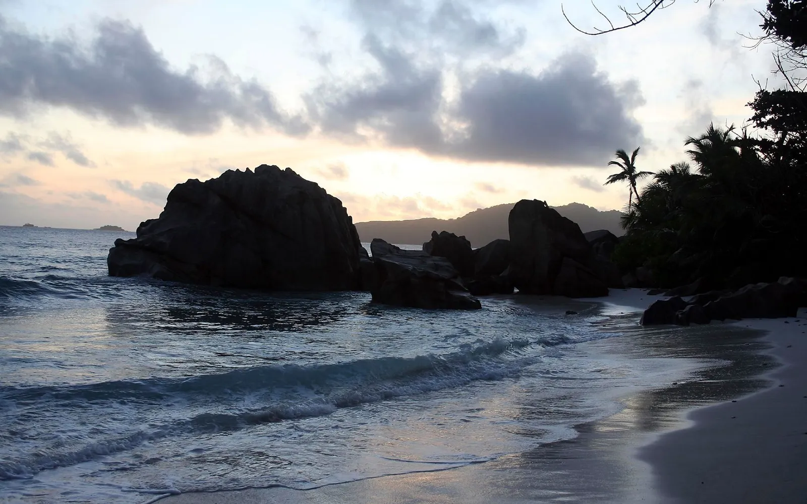 A serene beach scene with large granite boulders, gentle waves, and a cloudy sky at dusk.