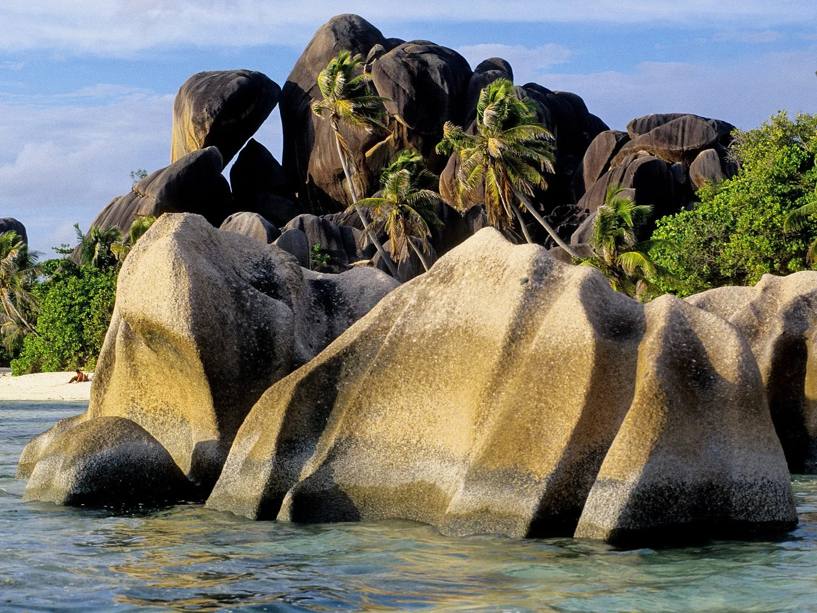 Granite boulders emerge from clear ocean water with palm trees in the background.