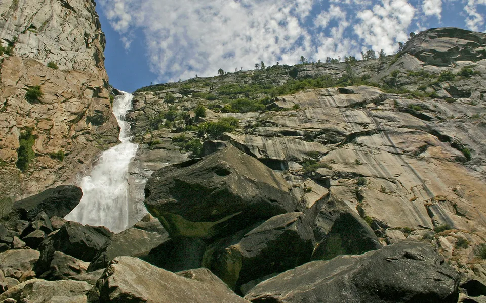 A tall waterfall cascades down a sheer granite cliff face with scattered clouds in the blue sky above.