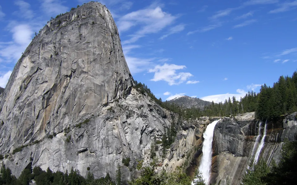 A tall waterfall plunges down a sheer granite cliff face, surrounded by pine trees under a blue sky with scattered clouds.