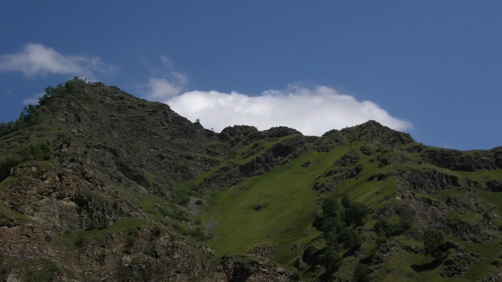 A view of rugged, green mountain slopes under a bright blue sky with scattered white clouds.