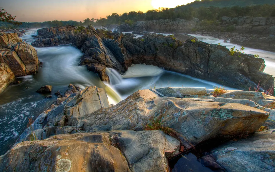 A wide river with rocky outcrops and smooth, flowing water, illuminated by the soft light of sunrise.