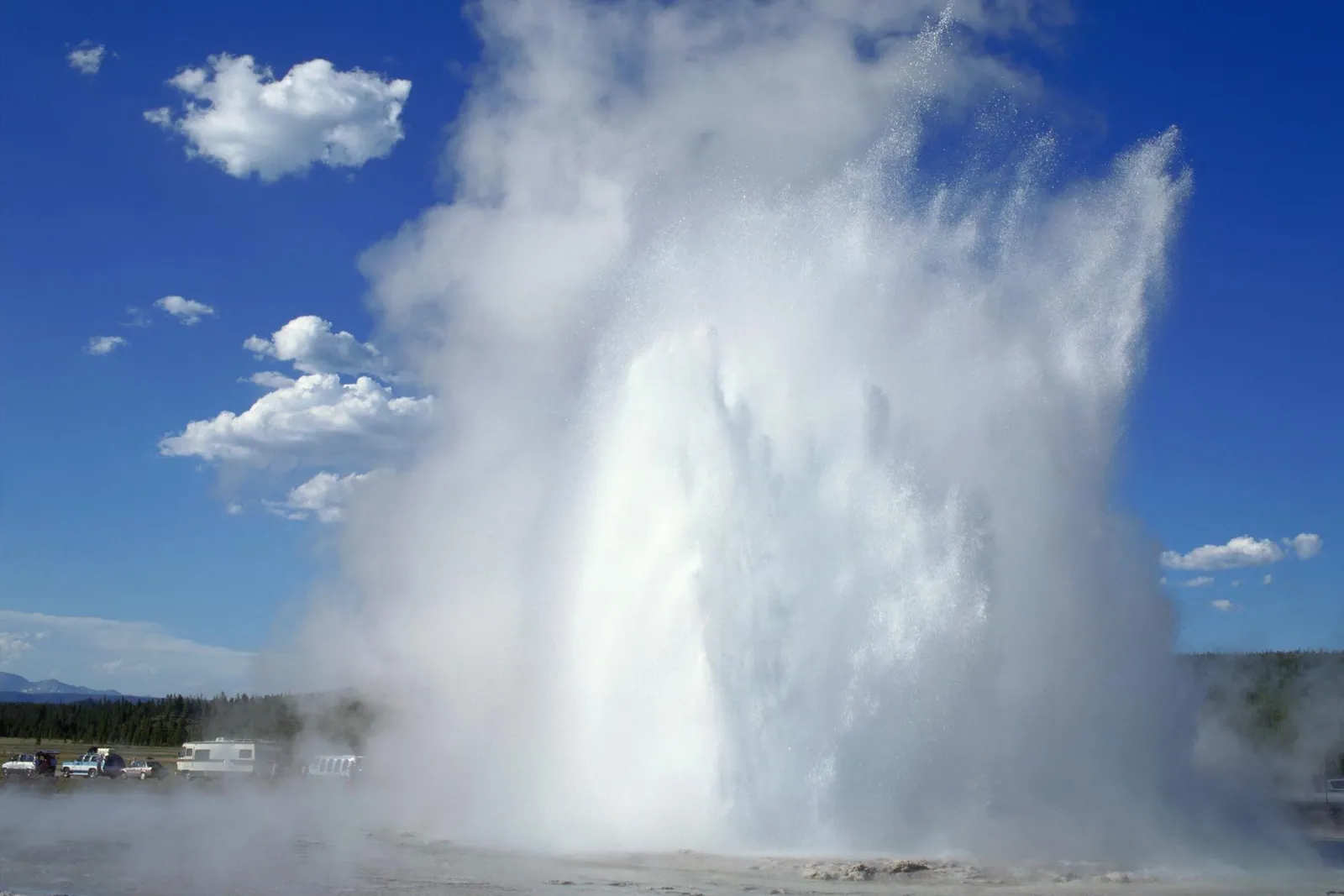 A powerful geyser erupts, sending a massive plume of water and steam high into a bright blue sky with scattered clouds.
