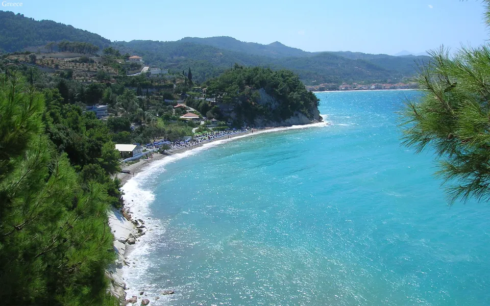 A scenic view of a Greek coastline with a beach, turquoise sea, and green hills dotted with houses.