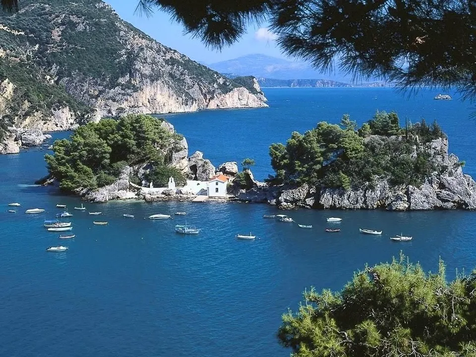 A small white village sits on a rocky island covered in trees, surrounded by blue water and dotted with boats.
