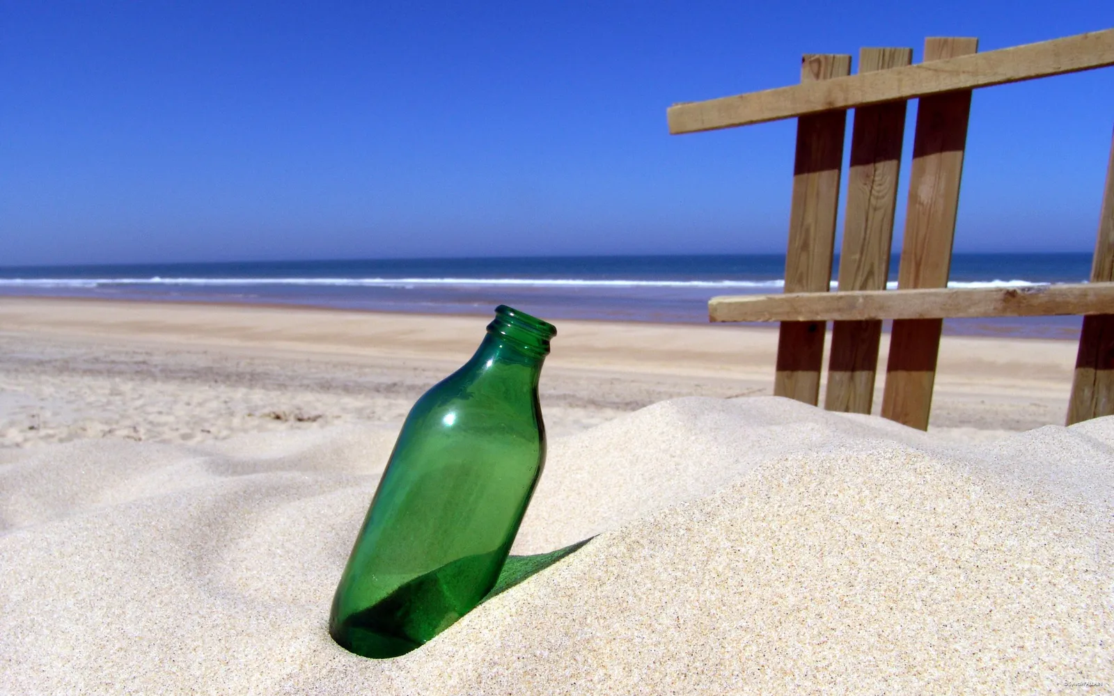A green glass bottle is half-buried in the sand on a beach, with the ocean and sky in the background.