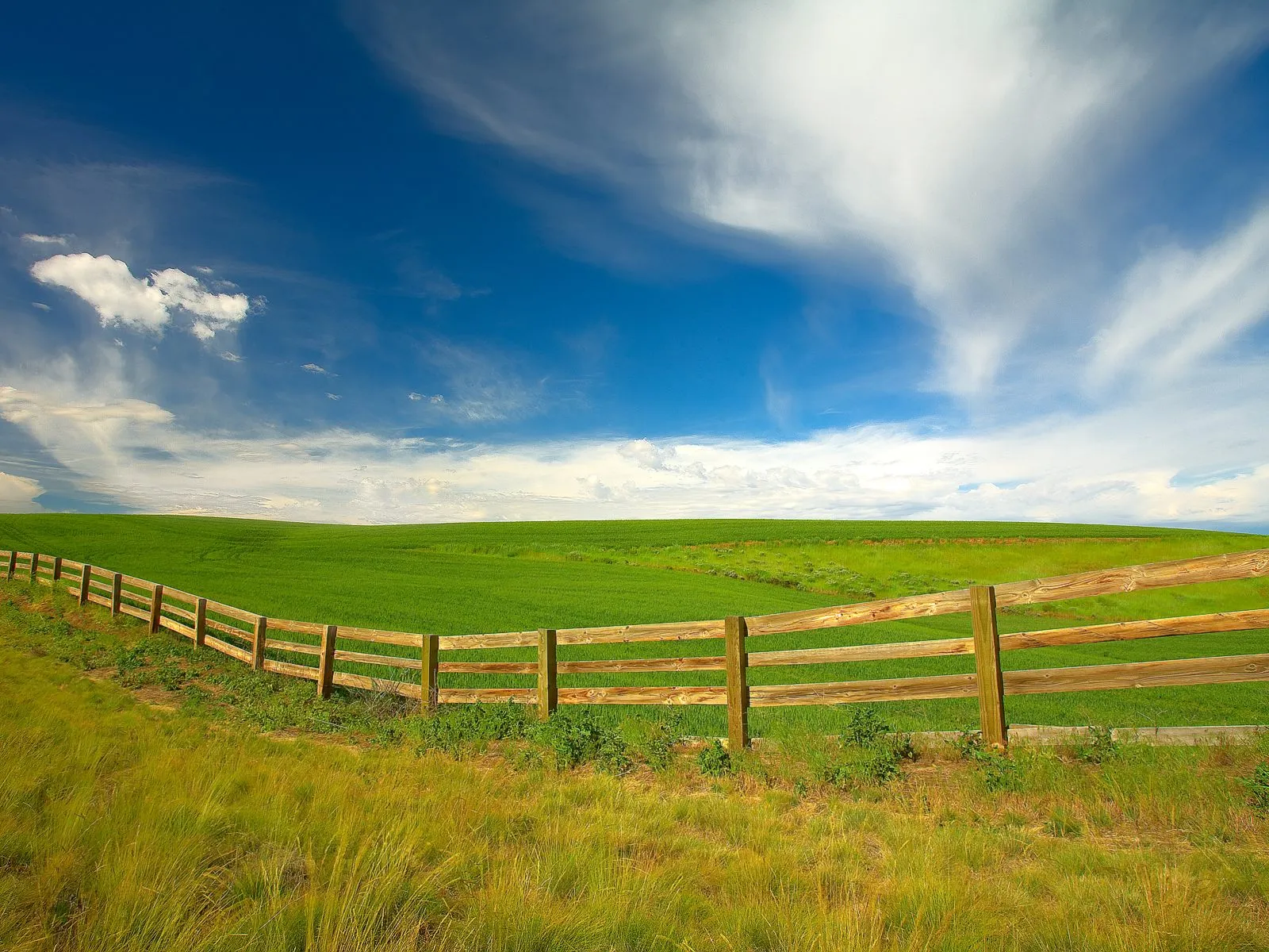A wooden fence runs along a grassy hill overlooking a vibrant green field under a blue sky with wispy clouds.
