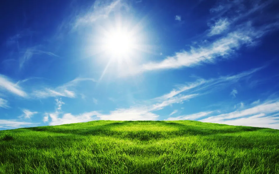 A bright sun shines in a blue sky with wispy clouds over a lush green grassy hill.