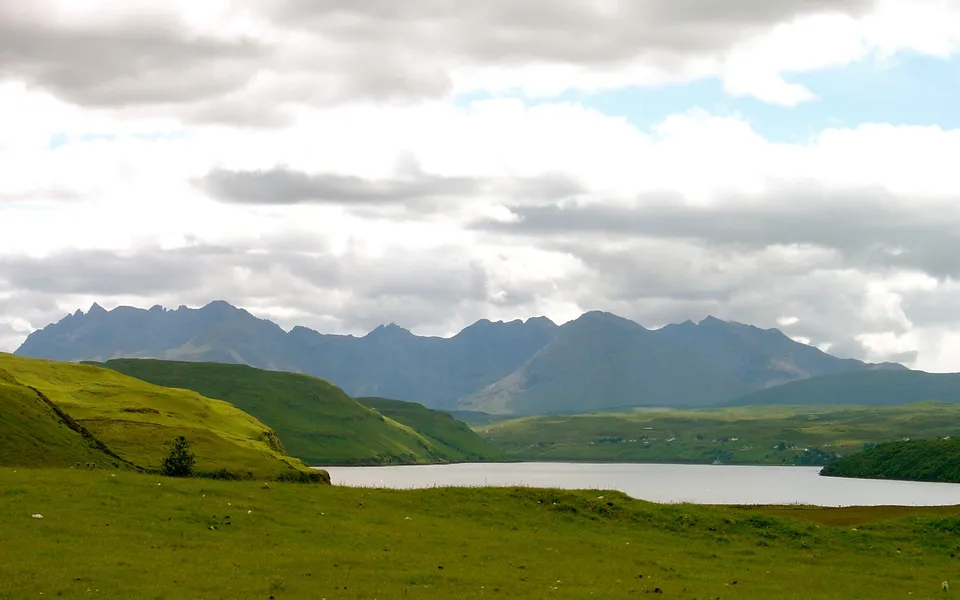 A wide shot of rolling green hills leading to a calm body of water with a dramatic mountain range in the background under a cloudy sky.