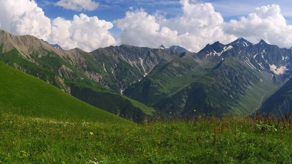 A panoramic view of a lush green mountain valley with dramatic peaks and a blue sky with white clouds.