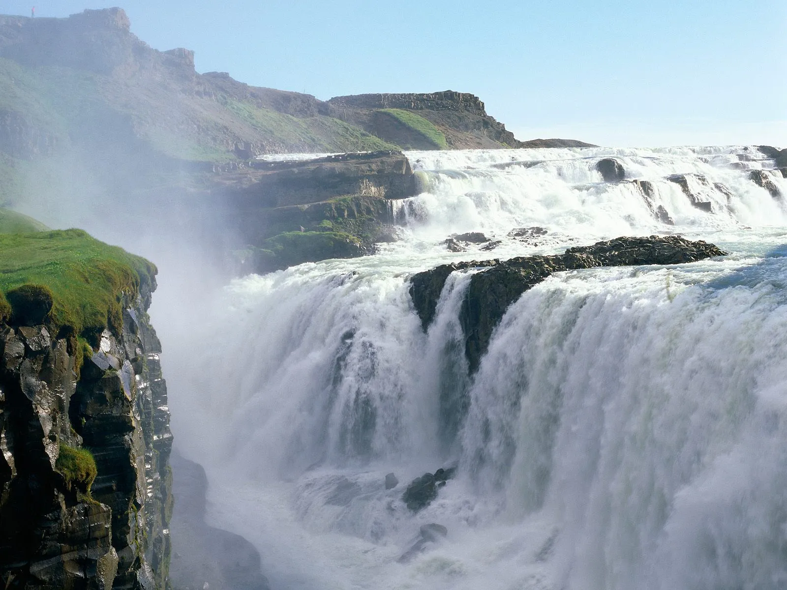 A wide, powerful waterfall cascades over multiple tiers of rock under a clear blue sky.