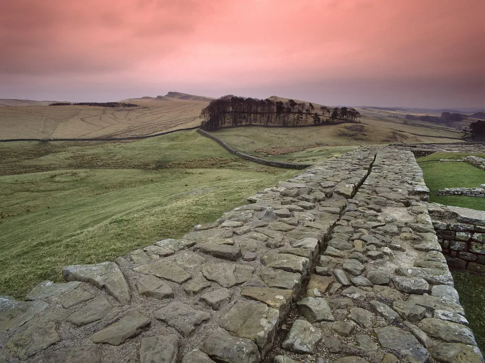A stone wall stretches into the distance across rolling green hills under a dramatic pink sky.