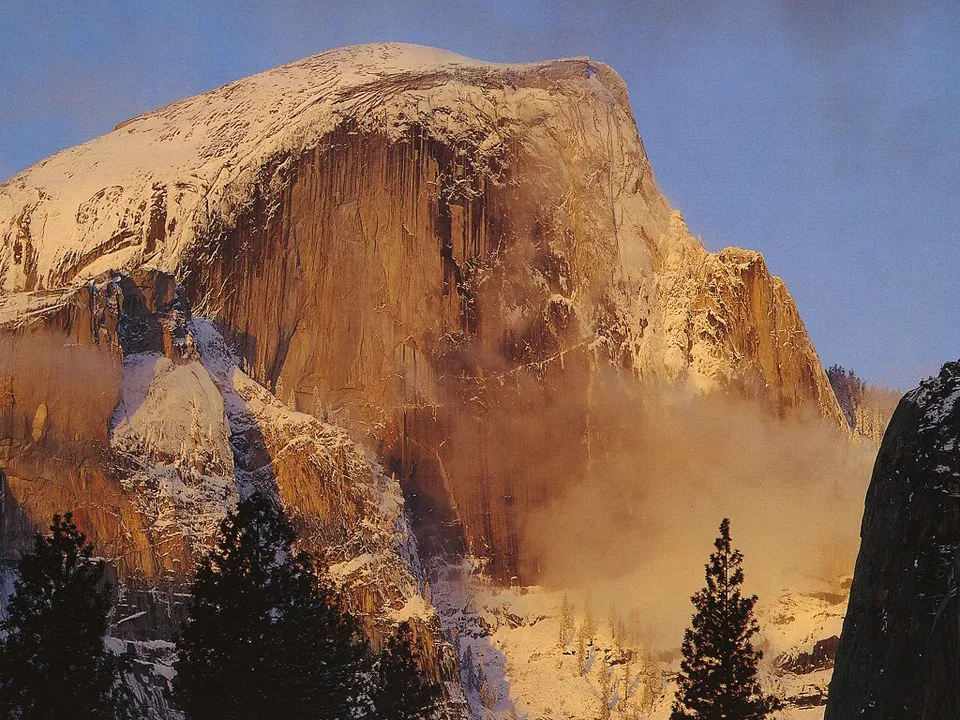A majestic snow-covered mountain peak, Half Dome, glows in the warm light of sunrise with clouds swirling around its base.