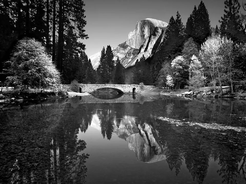 A stone bridge crosses a calm river reflecting Half Dome mountain and surrounding pine trees.