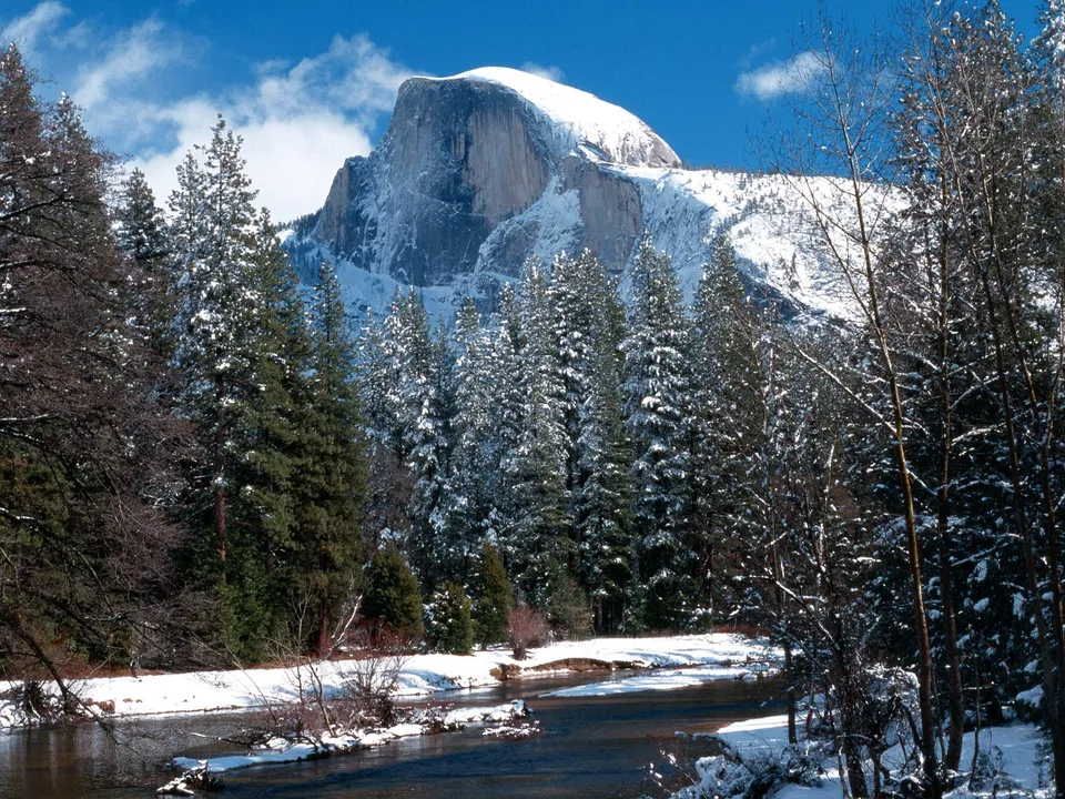 A majestic snow-covered mountain peak, Half Dome, rises above a forest of snow-laden pine trees and a winding river.