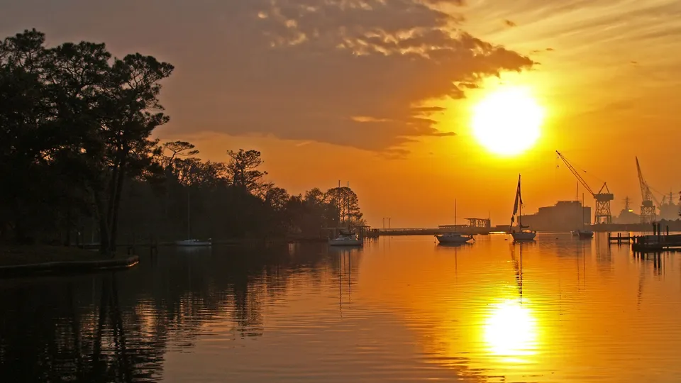 A bright sun rises over a calm harbor, casting golden light on the water and silhouetting trees and boats.