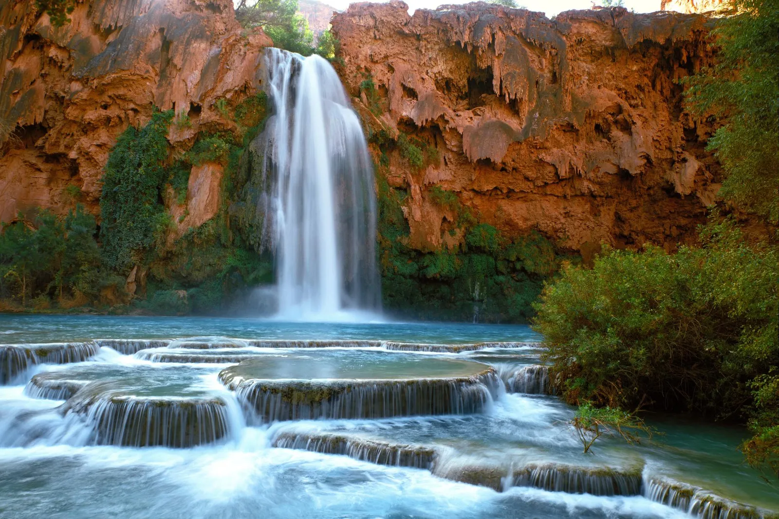 A wide waterfall cascades down a red rock cliff into turquoise pools with smaller tiered waterfalls in the foreground.