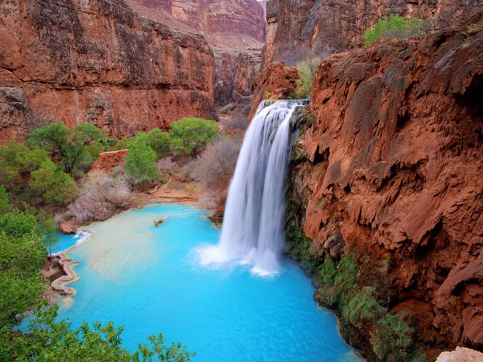 A wide waterfall cascades into a vibrant turquoise pool surrounded by red rock cliffs and green foliage.