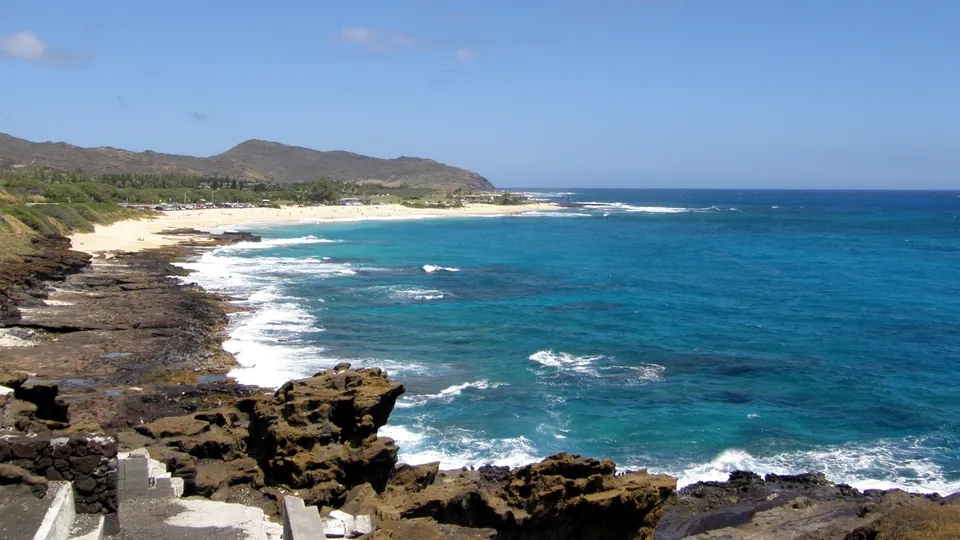 A view of a rocky coastline with turquoise ocean waves crashing against the shore, leading to a sandy beach and distant mountains.