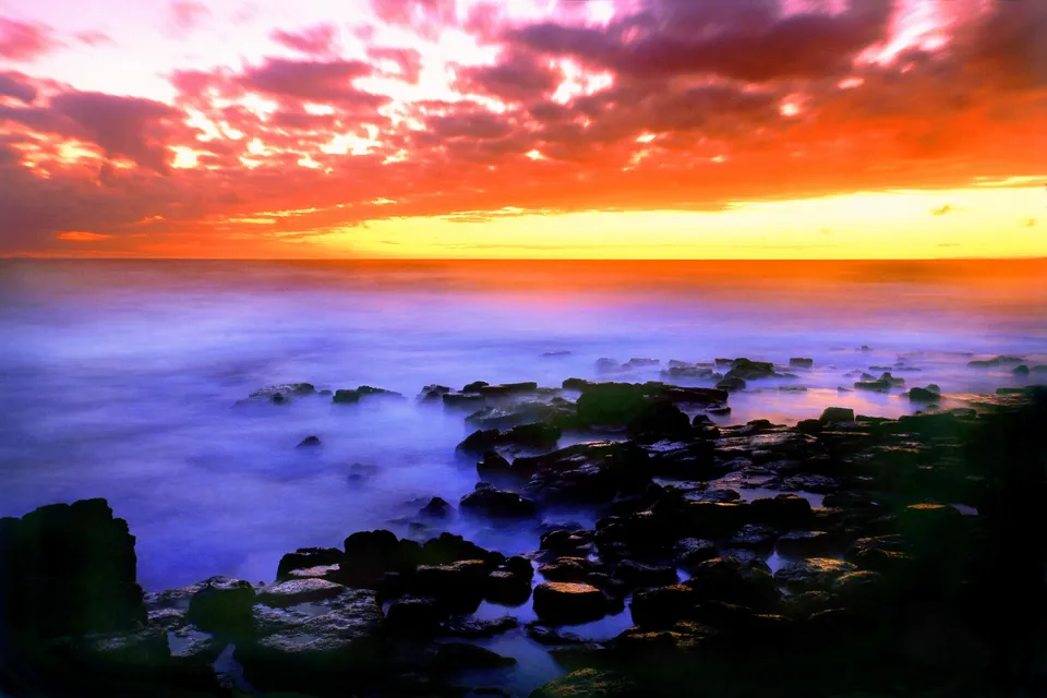 A dramatic sunset with fiery orange and pink clouds over a calm, misty ocean with dark rocks in the foreground.