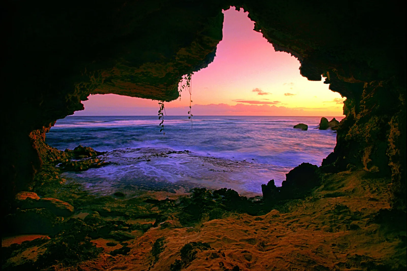 View from inside a dark sea cave looking out at a colorful sunset over the ocean.