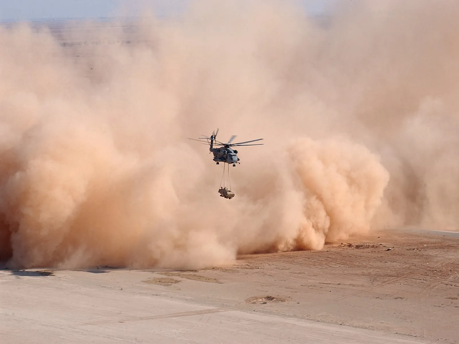 A military helicopter flies through a large, billowing dust storm in a desert landscape, carrying a load.