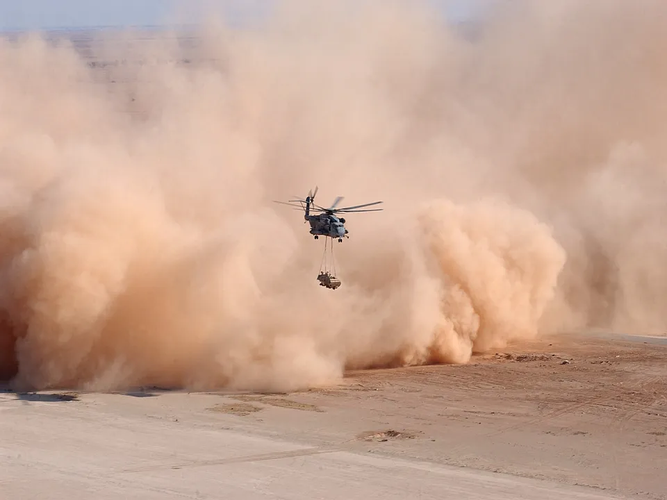 A military helicopter flies through a large, billowing dust storm in a desert landscape, carrying a load.