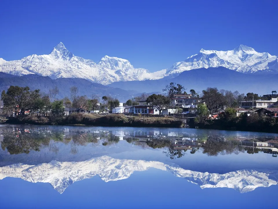 Snow-capped Himalayan mountains reflected perfectly in a calm lake with a town in the foreground.